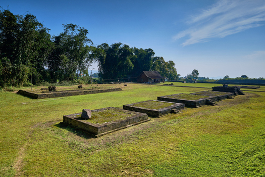 View of the ancient stone foundations of Situs Liyangan archaeological site surrounded by green grass and bamboo trees under a clear blue sky in Temanggung Central Java Indonesia.