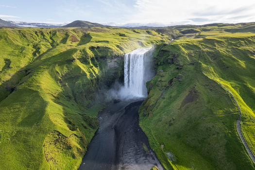 Aerial view of Skogafoss waterfall cascading down a steep cliff into a river surrounded by lush green hills and mountains Rangarthing eystra Iceland.