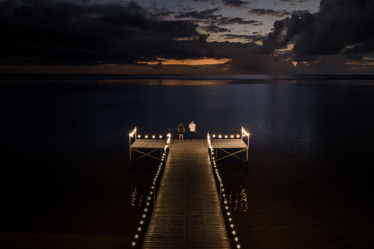 Aerial view of a wooden pier decorated with glowing string lights and two people standing at the end under a dark cloudy sky at dusk in Moka District Mauritius. Aerial view of a wooden pier decorated with glowing string lights and two people standing at the end under a dark cloudy sky at dusk in Moka District Mauritius.
