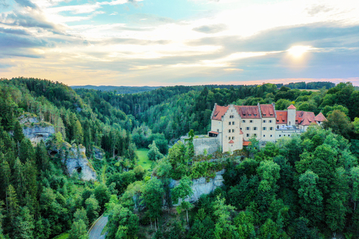 Aerial view of Burg Rabenstein perched on a rocky cliff surrounded by dense green forests under a soft sunset sky in Ahorntal Bayern Germany.