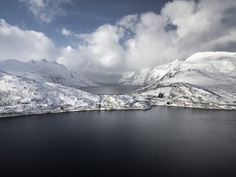 Aerial view of Grotfjord surrounded by snow covered mountains and a winding coastal road under a dramatic cloudy sky Tromvik Troms Norway. Aerial view of Grotfjord surrounded by snow covered mountains and a winding coastal road under a dramatic cloudy sky Tromvik Troms Norway.