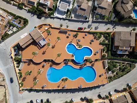 Aerial view of a residential complex with two interconnected blue swimming pools a terracotta colored deck palm trees and nearby villas in Moraira Valencian Community Spain.