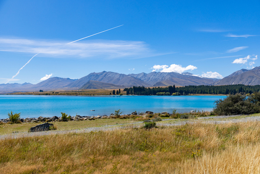 View of turquoise lake water meeting golden grasslands under a vast blue sky streaked with contrails framed by majestic mountains Lake Tekapo New Zealand. View of turquoise lake water meeting golden grasslands under a vast blue sky streaked with contrails framed by majestic mountains Lake Tekapo New Zealand.