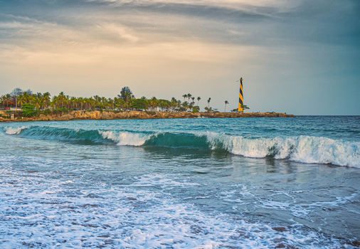 View of the Sans Souci lighthouse standing on the rocky coastline with palm trees and crashing waves under a cloudy sky in Santo Domingo Este Santo Domingo Provinc Dominican Republic.