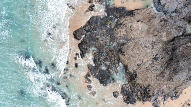 Aerial view of turquoise waves crashing against the dark jagged rocks and sandy shore of Constantine Bay in Padstow Cornwall United Kingdom. Aerial view of turquoise waves crashing against the dark jagged rocks and sandy shore of Constantine Bay in Padstow Cornwall United Kingdom.