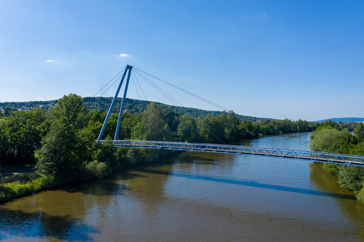 Aerial view of the blue cable stayed pedestrian bridge at Sportinsel Forchheim crossing the Regnitz river surrounded by dense green trees and hills Forchheim Bayern Germany.