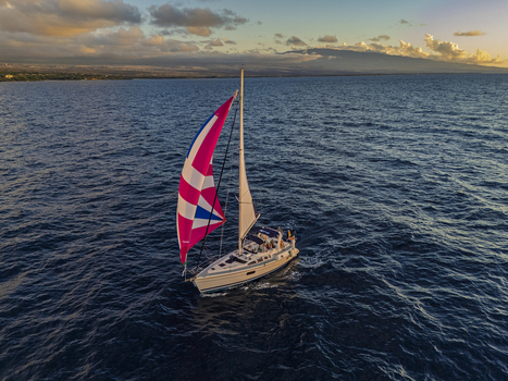 Aerial view of a sailboat with a colorful pink spinnaker sailing on the blue ocean near the coastline of Kawaihae Harbor under a golden sunset sky in Waimea Hawaii United States. Aerial view of a sailboat with a colorful pink spinnaker sailing on the blue ocean near the coastline of Kawaihae Harbor under a golden sunset sky in Waimea Hawaii United States.