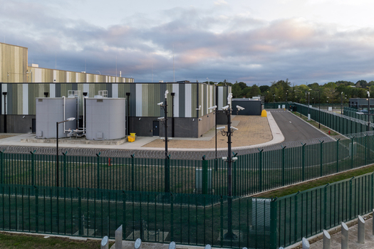 Aerial view of a data center with industrial buildings large storage tanks and a secure perimeter fence with surveillance cameras under a cloudy sky in the United Kingdom. Aerial view of a data center with industrial buildings large storage tanks and a secure perimeter fence with surveillance cameras under a cloudy sky in the United Kingdom.