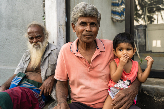Sri Lanka 15 January 2026 View of an elderly man with grey hair holding a young child in his arms while another man with a long white beard sits in the background.