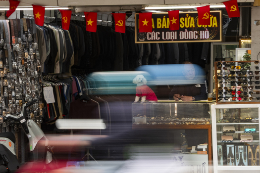 Hanoi Vietnam 11 November 2025 View of a shop in Hanoi with a man behind a glass counter and a small dog in a red shirt surrounded by hanging clothes and motion blurred traffic.