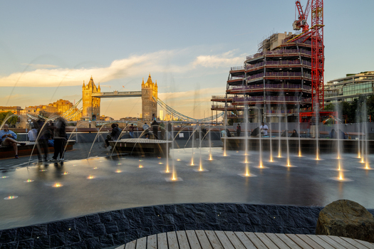 View of The Scoop fountain with illuminated water jets Tower Bridge in the background and a construction site with a crane in London England United Kingdom. View of The Scoop fountain with illuminated water jets Tower Bridge in the background and a construction site with a crane in London England United Kingdom.