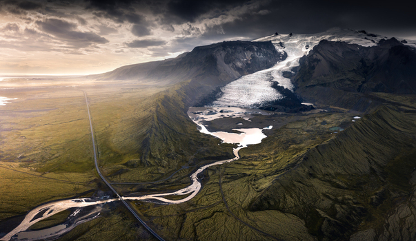 Aerial view of a massive glacier tongue flowing into a lagoon next to a long straight road under a dramatic cloudy sky in Iceland.