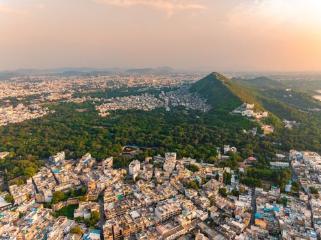Aerial view of the dense urban landscape of Udaipur featuring a large green forest a prominent hill and buildings bathed in the warm light of the golden hour in Udaipur Rajasthan India.