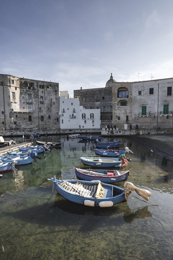 View of traditional blue fishing boats moored in the clear waters of the old harbor with people on the quay and historic stone buildings in Polignano a Mare Apulia Italy.