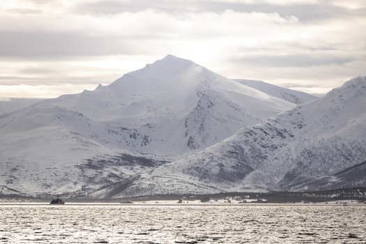 View of snow covered mountain peaks overlooking a fjord with a small ship on the water under a cloudy sky Tromso Troms Norway.