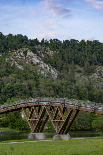 View of the arched wooden bridge over the river with lush green forests and rocky cliffs under a soft sky in Essing Bayern Germany.