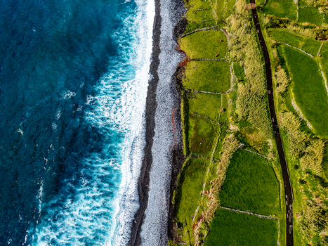 Aerial view of the contrasting blues of the ocean meeting the dark volcanic beach and bright green fields Faj&atilde; Grande Azores Portugal.