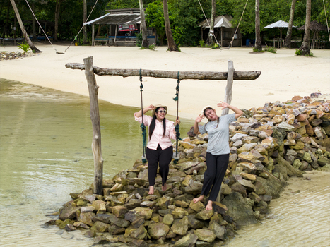 Aerial view of two women enjoying a swing along Bobby Beach where turquoise waters meet the sandy shore under the shade of swaying palms Karimunjawa Central Java Indonesia.