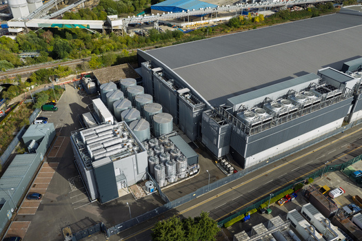 Aerial view of a large data center facility featuring industrial cooling towers metal storage tanks and a secure parking area in the United Kingdom. Aerial view of a large data center facility featuring industrial cooling towers metal storage tanks and a secure parking area in the United Kingdom.