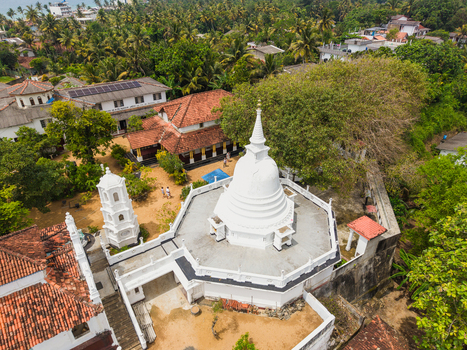 Aerial view of the white stupa of Kumarakanda Kumara Maha Viharaya surrounded by traditional buildings and lush palm trees in Hikkaduwa Southern Province Sri Lanka. Aerial view of the white stupa of Kumarakanda Kumara Maha Viharaya surrounded by traditional buildings and lush palm trees in Hikkaduwa Southern Province Sri Lanka.
