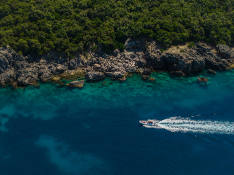 Aerial view of Aquarium Beach featuring a white motorboat cruising through turquoise waters along a rocky coastline with lush green forest in Jala Vlore County Albania. Aerial view of Aquarium Beach featuring a white motorboat cruising through turquoise waters along a rocky coastline with lush green forest in Jala Vlore County Albania.
