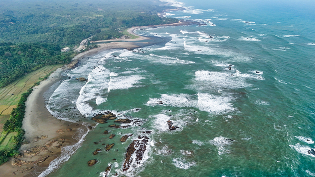 Aerial view of a rugged coastline with a sandy beach rocky outcrops in the turquoise sea crashing waves and lush green tropical vegetation in Indonesia. Aerial view of a rugged coastline with a sandy beach rocky outcrops in the turquoise sea crashing waves and lush green tropical vegetation in Indonesia.