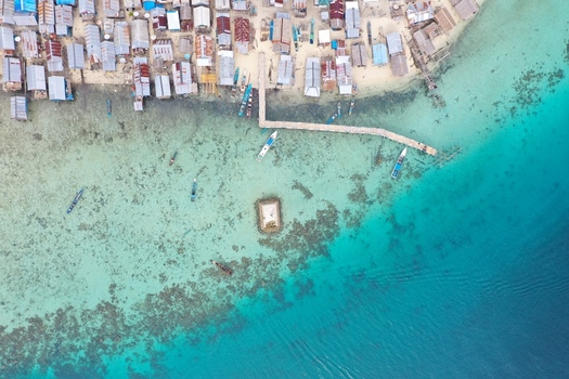 Aerial view of Pulau Maginti featuring a coastal village with stilt houses a wooden pier extending into the turquoise sea and small boats anchored in clear coral rich waters Maginti South East Sulawesi Indonesia.