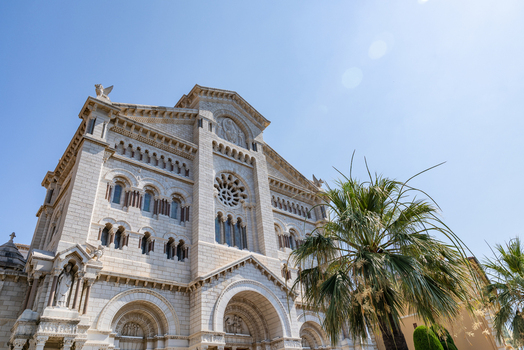 View of the Saint Nicholas Cathedral with its ornate white stone facade rose window and a palm tree in the foreground under a clear blue sky in Monaco City Monaco. View of the Saint Nicholas Cathedral with its ornate white stone facade rose window and a palm tree in the foreground under a clear blue sky in Monaco City Monaco.