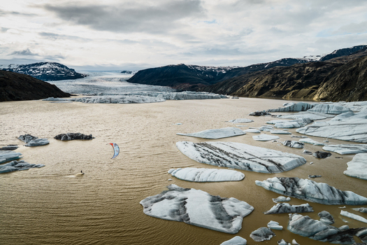 Aerial view of a kiteboarder gliding across the glacial waters of Vatnajokull surrounded by floating icebergs and dramatic mountain landscapes in Iceland.