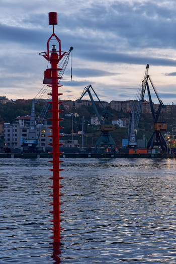 Pasai Donibane Spain 09 March 2026 View of a red navigation beacon in the water with industrial harbor cranes and coastal buildings under a cloudy twilight sky.