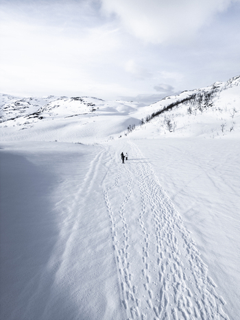 Aerial view of snowy mountain slopes with a footprint covered path and people walking in the distance under a bright sky in Kvaloya Troms Norway. Aerial view of snowy mountain slopes with a footprint covered path and people walking in the distance under a bright sky in Kvaloya Troms Norway.
