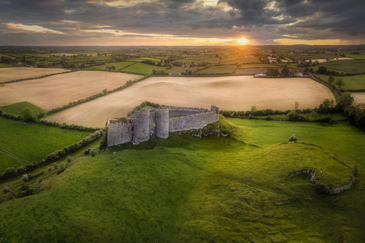 Aerial view of Castle Roche ruins on a grassy hill overlooking golden fields at sunset in County Louth Ireland.