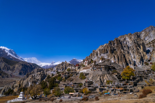 Aerial view of a traditional mountain village with stone houses and a monastery nestled among rugged rocky cliffs and snow capped Himalayan peaks under a clear blue sky in Nepal.