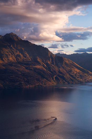 Aerial view of Lake Wakatipu with a small boat leaving a wake on the calm water surrounded by dramatic mountains under a golden hour sky in Queenstown Otago Region New Zealand. Aerial view of Lake Wakatipu with a small boat leaving a wake on the calm water surrounded by dramatic mountains under a golden hour sky in Queenstown Otago Region New Zealand.
