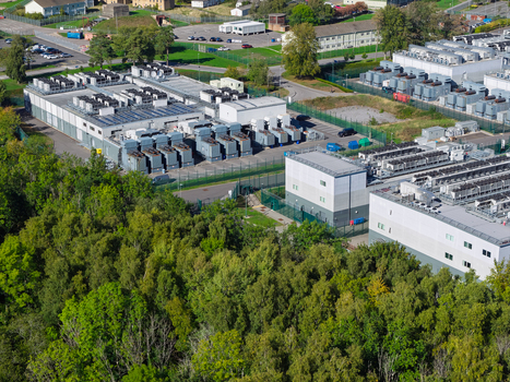 Aerial view of a modern data center facility with multiple industrial buildings rooftop cooling units and fenced perimeter surrounded by dense green forest in the United Kingdom.