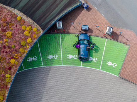 Aerial view of a blue electric vehicle parked in a designated green charging spot with a person standing beside it next to a building with a green roof in Beilen Drenthe Netherlands.