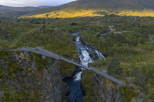 Aerial view of the Voringfossen waterfall and the modern pedestrian bridge spanning the deep rocky canyon surrounded by autumn foliage in Eidfjord Municipality Vestland Norway.