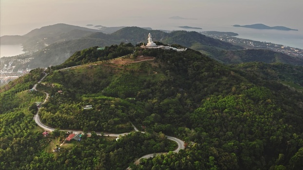 Aerial view of the Big Buddha statue perched atop a lush green hill surrounded by dense tropical forest and winding roads overlooking the coastline in Phuket Phuket Thailand. Aerial view of the Big Buddha statue perched atop a lush green hill surrounded by dense tropical forest and winding roads overlooking the coastline in Phuket Phuket Thailand.