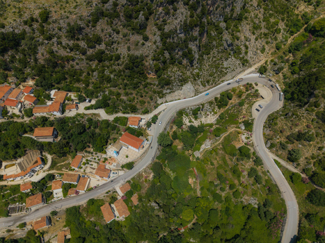 Aerial view of a winding road passing through a hillside village with traditional red roofed houses nestled among green hills and rocky slopes Dhermi Vlore County Albania. Aerial view of a winding road passing through a hillside village with traditional red roofed houses nestled among green hills and rocky slopes Dhermi Vlore County Albania.