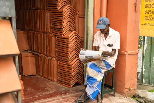 Sri Lanka 12 February 2026 View of an elderly man reading a newspaper while sitting on a stool next to stacks of terracotta roof tiles outside a shop.