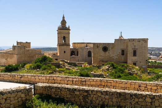 View of the historic stone church and bell tower surrounded by ancient stone walls and green vegetation under a clear blue sky in Zebbug Gozo Region Malta. View of the historic stone church and bell tower surrounded by ancient stone walls and green vegetation under a clear blue sky in Zebbug Gozo Region Malta.