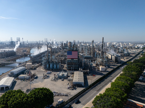Aerial view of an industrial oil refinery featuring a large American flag on a central structure surrounded by storage tanks and smoke stacks in Carson California United States.