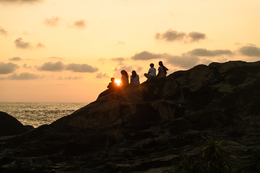 Sri Lanka 25 January 2026 View of a group of people sitting on a rocky cliff by the ocean during a vibrant sunset with golden light and scattered clouds.