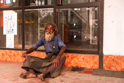 Sri Lanka 14 January 2026 View of an elderly man with a long beard and messy hair sitting on the ground in front of a shop window with posters.