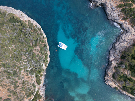Aerial view of Cala Sa Nau bay with a white catamaran anchored in turquoise waters between rocky cliffs in Felanitx Balearic Islands Spain. Aerial view of Cala Sa Nau bay with a white catamaran anchored in turquoise waters between rocky cliffs in Felanitx Balearic Islands Spain.