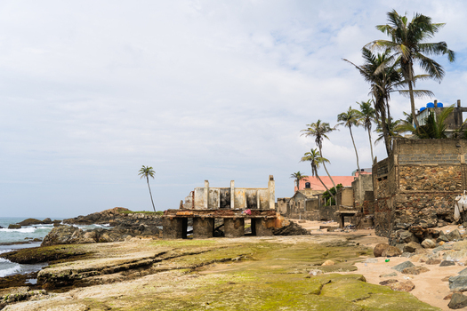 View of ruined coastal buildings and palm trees along the rocky shoreline under a cloudy sky in Galle Southern Province Sri Lanka.
