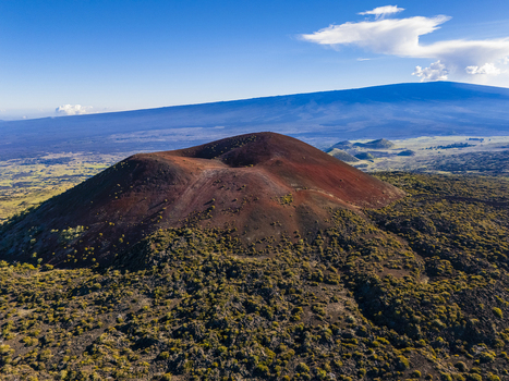 Aerial view of the Mauna Kea volcanic cinder cone with reddish soil surrounded by green shrubs and lava fields under a clear blue sky Hawaii County Hawaii United States.