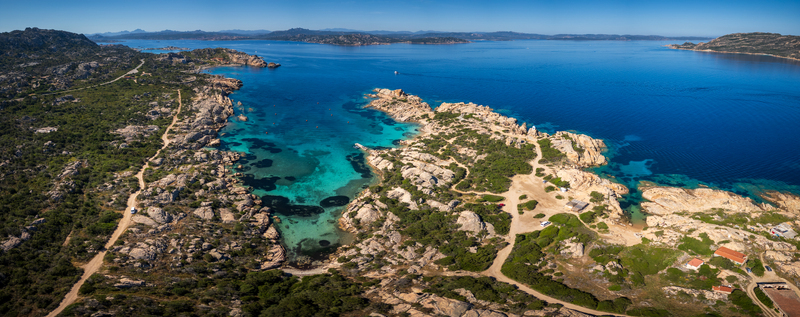 Aerial view of the rocky coastline of Punta Carlotto with turquoise sea water and winding dirt paths under a clear blue sky in La Maddalena Sardinia Italy. Aerial view of the rocky coastline of Punta Carlotto with turquoise sea water and winding dirt paths under a clear blue sky in La Maddalena Sardinia Italy.