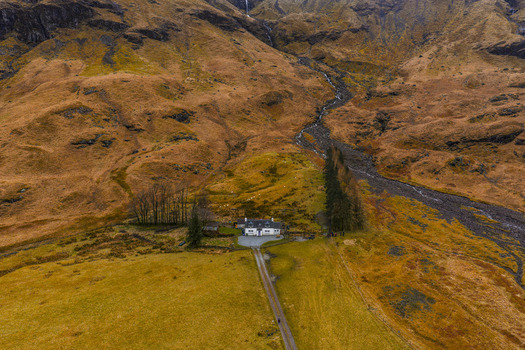 Aerial view of a white cottage at the base of rugged mountains with a winding stream and autumn colored slopes in Glencoe Scotland United Kingdom. Aerial view of a white cottage at the base of rugged mountains with a winding stream and autumn colored slopes in Glencoe Scotland United Kingdom.