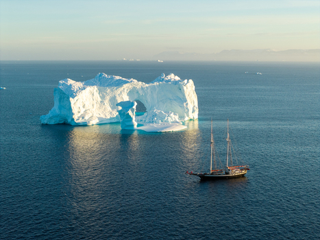 Aerial view of a large arch iceberg floating in the deep blue sea near a sailing ship in Greenland. Aerial view of a large arch iceberg floating in the deep blue sea near a sailing ship in Greenland.